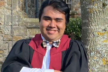 A male postgraduate scholar wearing University of St Andrews black and burgundy postgraduate gown and a bow tie, leaning against a tree